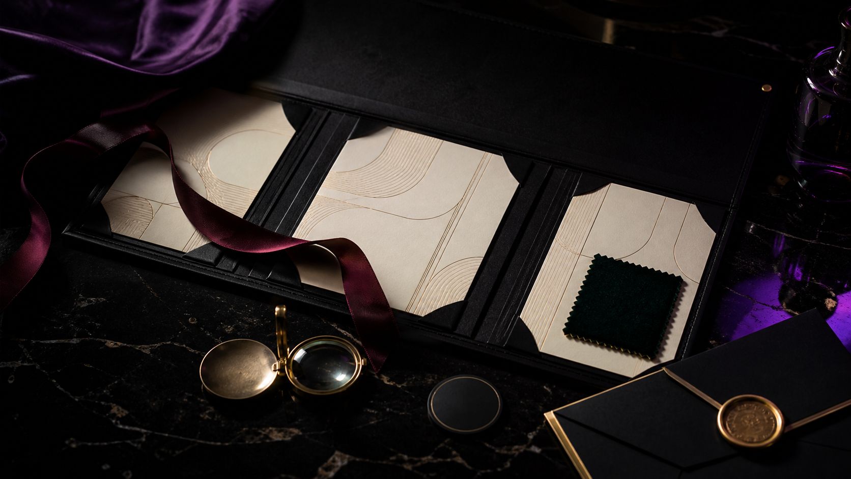 Blank cream term cards, emerald fabric, burgundy ribbon, brass loupe, and sealed envelope in a black folio